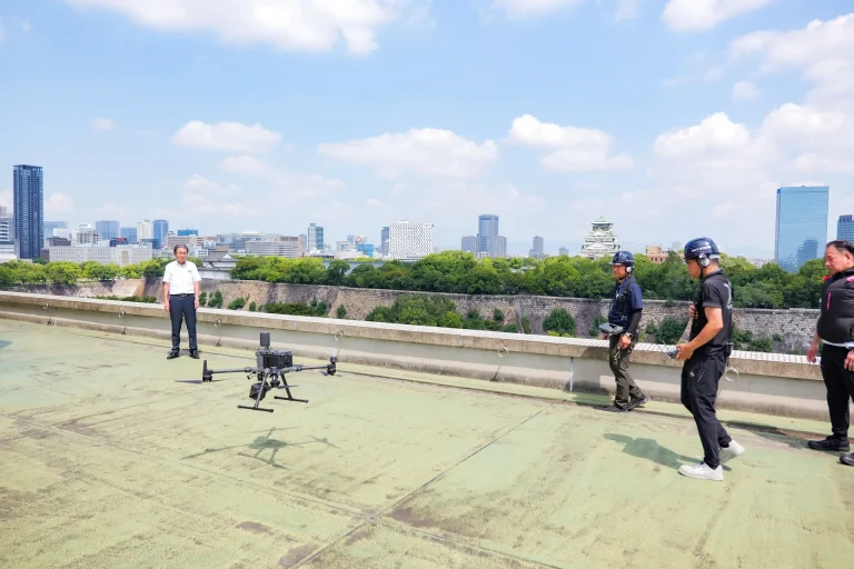 大阪市中央区のホテル屋上。ドローンの前に調査メンバーが立ち、背景に大阪城が見える
